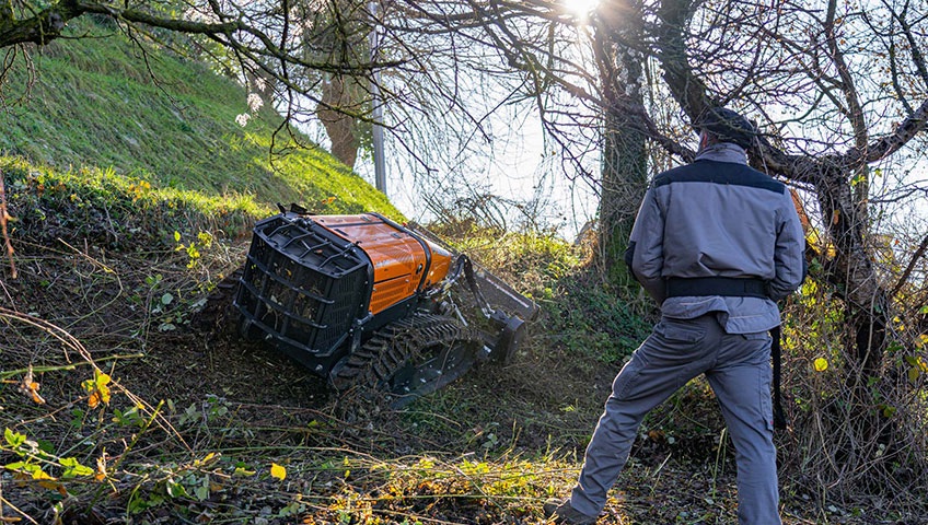 Operator watching a remote-controlled tracked mower clear brush on a steep slope